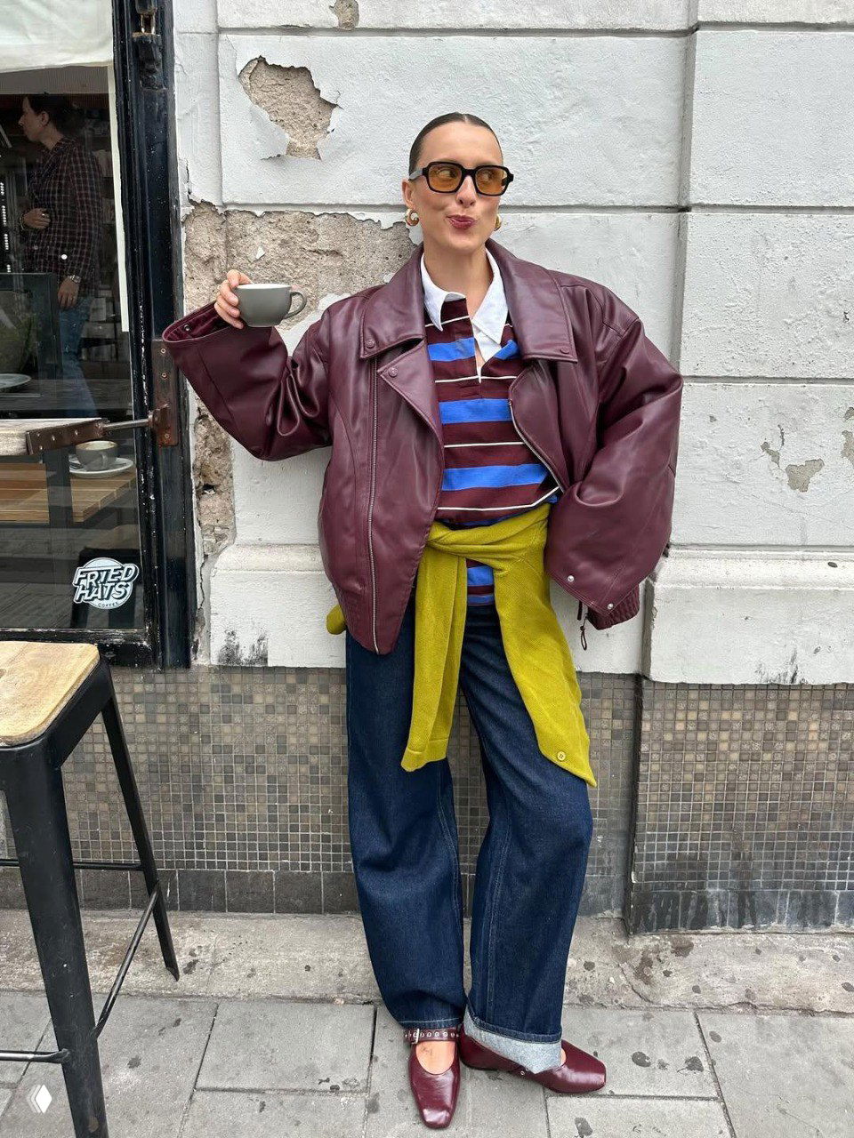 Стритстайл: женщина в бордовой кожаной куртке и оливковом кардигане, широкие джинсы и бордовые туфли у кафе, контрастное сочетание цветов в образе.