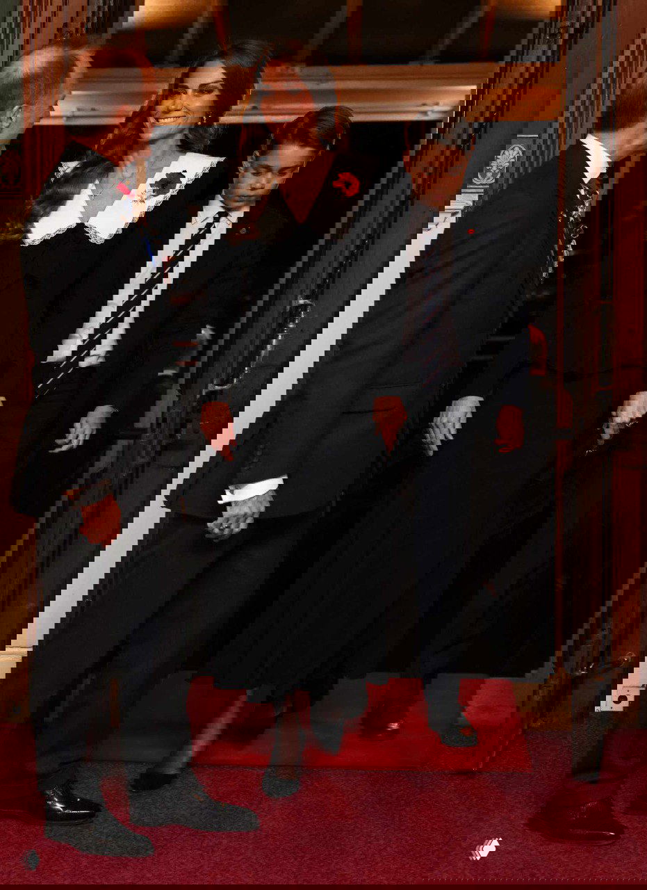 Женщина в чёрном платье с белым кружевным воротником и мальчик в тёмном костюме идут по красной дорожке в торжественном зале.