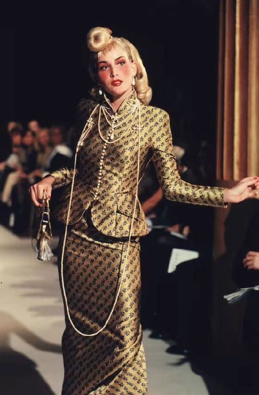 Модель в золотом костюме со сложным узором и длинной жемчужной нитью, прическа и макияж подчёркивают винтажную эстетику показа Dior осень 1998.