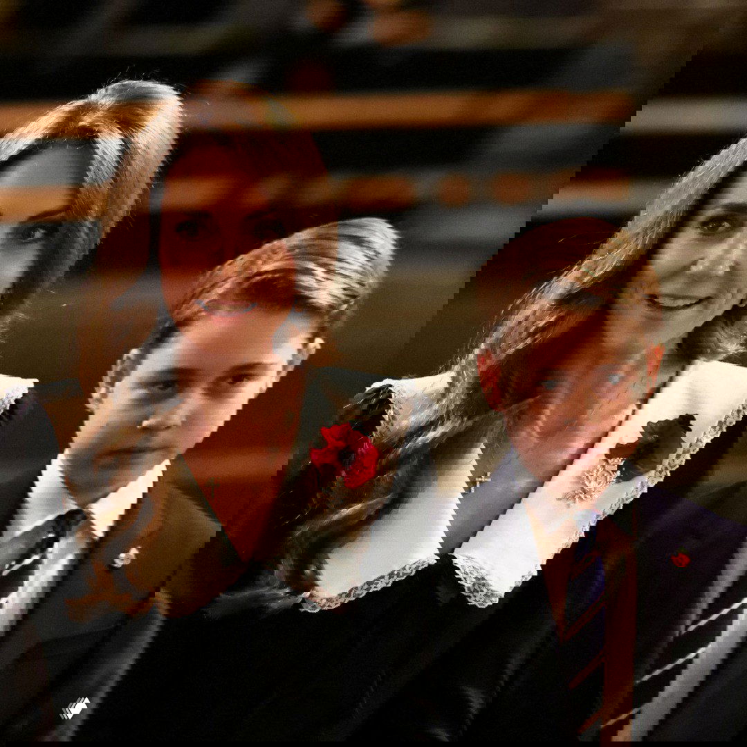 Крупный план женщины в чёрном платье с белым воротником рядом с мальчиком в тёмном пиджаке в интерьере концертного зала.