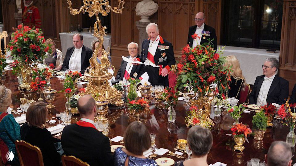 Вид на большой банкетный стол с сидящими гостями и королём, произносящим тост, в богато украшенном зале замка.