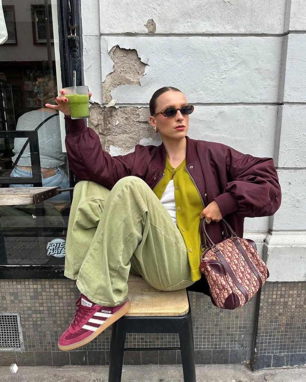 Уличная съёмка: бордовый бомбер, оливковые вельветовые брюки и бордовые кеды, модель сидит в кафе с напитком — игра контраста и фактур.