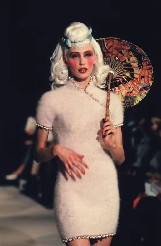 Модель в пушистом белом платье с декоративным зонтиком, ретро‑прическа и нежный макияж — кадр показательный для стилистики Dior осень 1998 на подиуме.