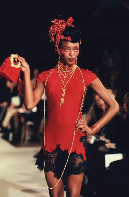 Модель в ярком красном платье с длинными золотыми бусами на подиуме Dior 1998, контрастный макияж и декоративный головной аксессуар создают драматичный образ.