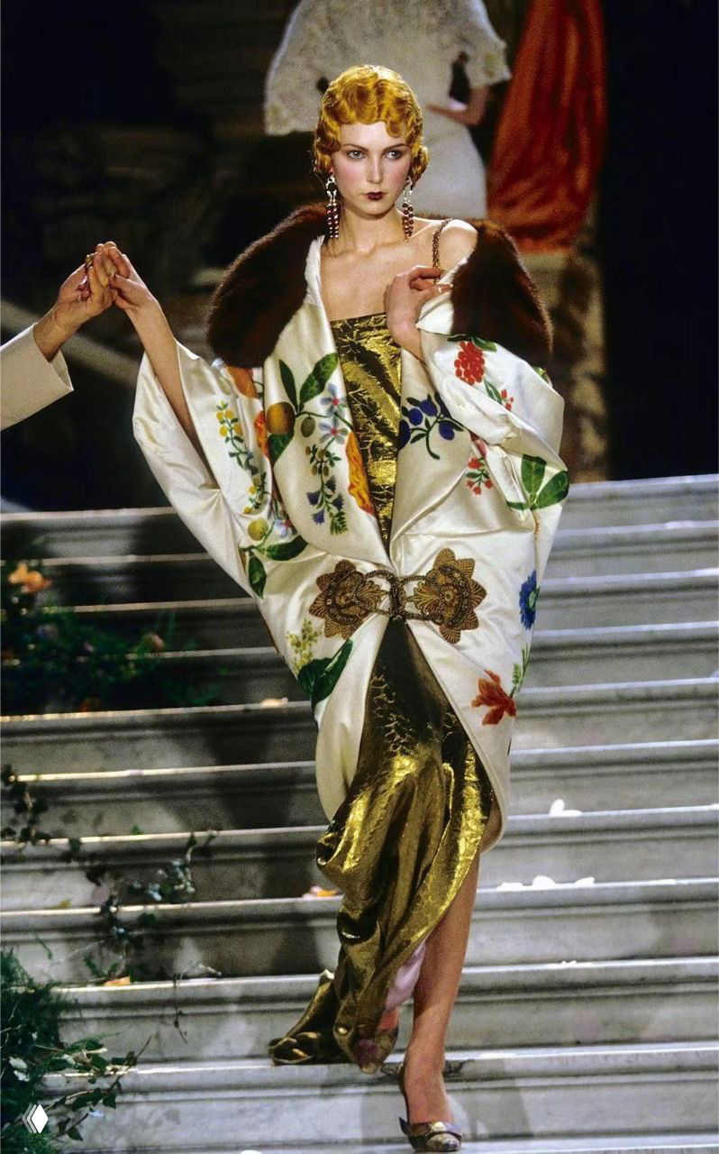 Модель в атласном платье с просторным вышитым плащом с цветочной аппликацией и меховой отделкой, образ в духе старинного гламура на показе Dior 1998.