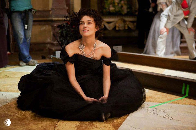 Героиня в том же чёрном платье в сидячей позе на съёмочной площадке; видны жемчужные украшения и детали верхней части наряда.