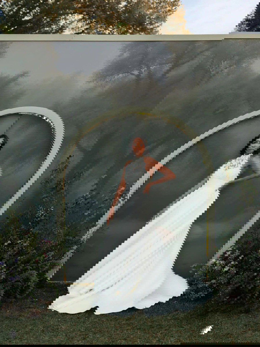 Винус стоит в элегантном белом платье у арочной декорации в саду — образ с высоким воротником и объёмной развевающейся юбкой.