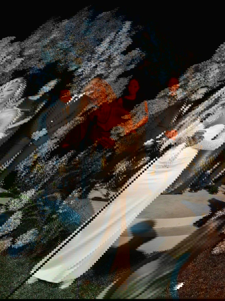 Невеста выступает с микрофоном в облегающем белом платье на ночном приёме; на заднем плане гости и цветочная инсталляция создают атмосферу.