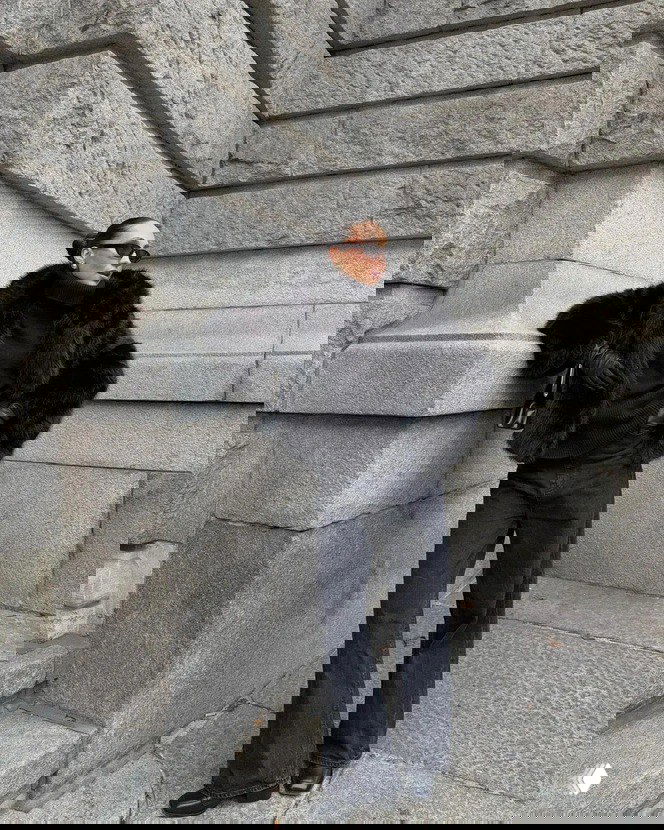 Модель в чёрной короткой шубе на фоне каменного здания, образ в монохроме с тёмными джинсами и перчатками, городской стритстайл