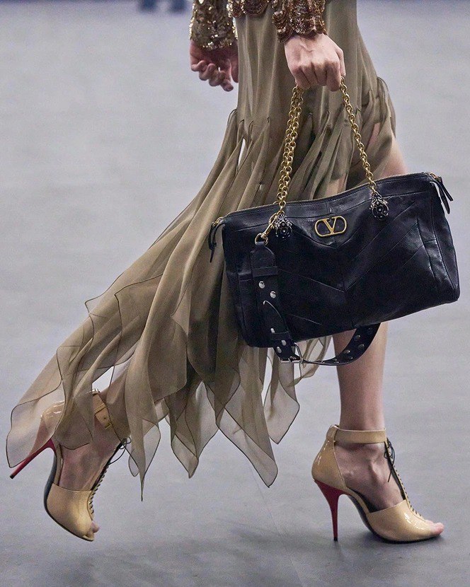Модель идёт по подиуму с чёрной сумкой Valentino Panthea: матовая кожа, V‑логотип и массивная золотая цепочка-ручка, сумка в движении образа на показе.