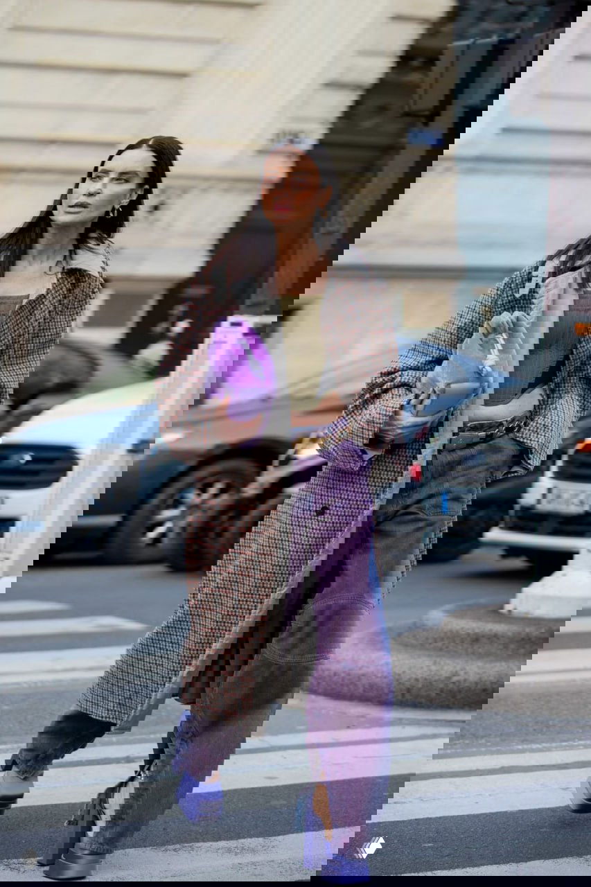 Женщина в сиренево-фиолетовых широких брюках, сиреневой сумке и клетчатом пальто идёт по городу — городской стритстайл с фиолетовыми акцентами.