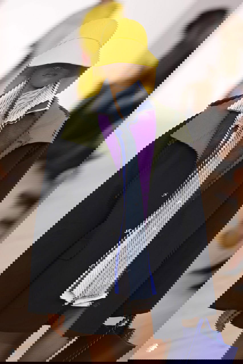 Модель на показе в многоуровневом образе: сиреневый стёганый жилет под тёмным плащом и ярко-жёлтая шляпа — сочетание цвета и фактуры на подиуме.