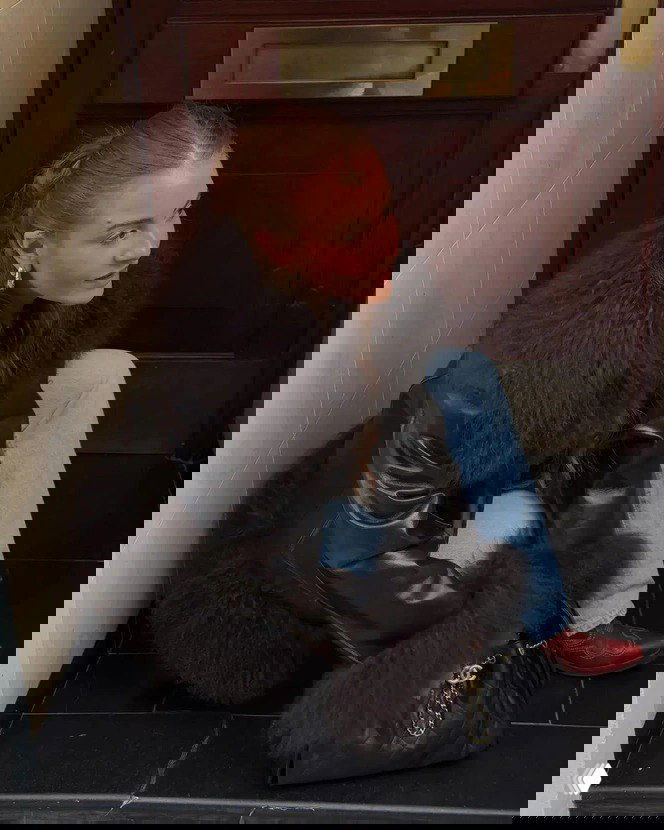 Женщина в кожаном пальто со меховыми вставками тёмного цвета сидит в дверном проёме, образ комбинирует кожу и пушистый мех