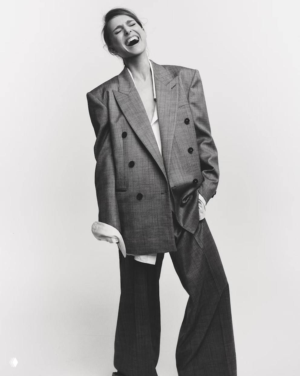 Чёрно-белый снимок в широком двубортном пиджаке и свободных брюках, артикулированная поза и выражение радости, классическая журнальная стилистика.