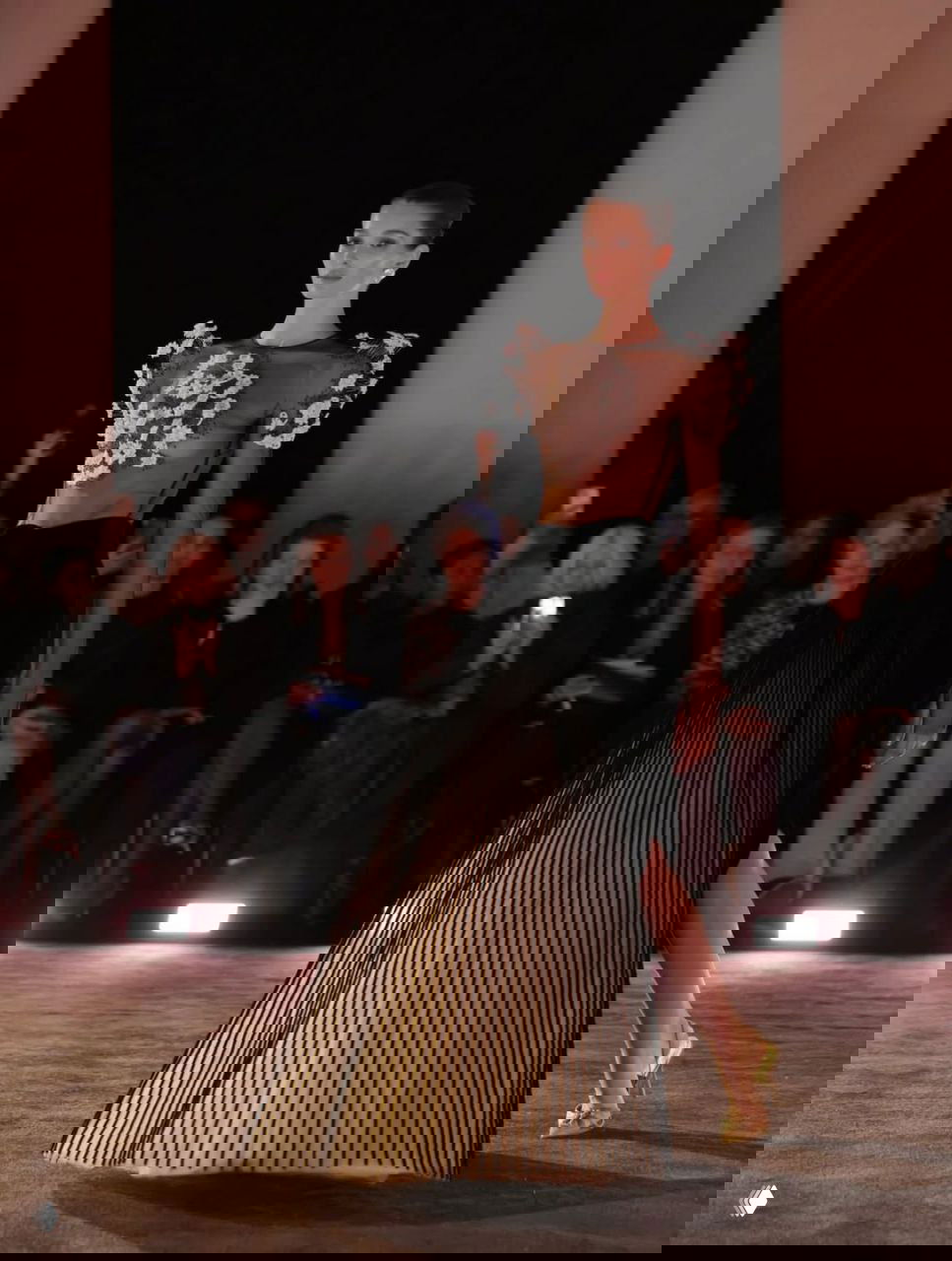 Фигура модели в прозрачном лифе с цветочными аппликациями и плиссированной юбкой, световой дизайн подчеркивает текстуру ткани на показе.