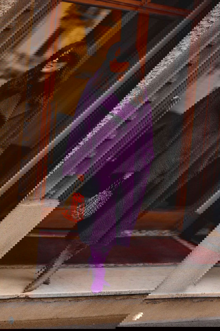 Женщина в длинном пушистом пурпурном пальто и сиреневых ботильонах выходит из двери отеля — роскошный текстурный образ для холодного сезона.