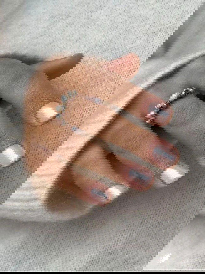 Рука в светлом свитере с аккуратными овальными ногтями в персиково-розовом перламутровом тоне и украшением-кольцом, холодная зимняя палитра.