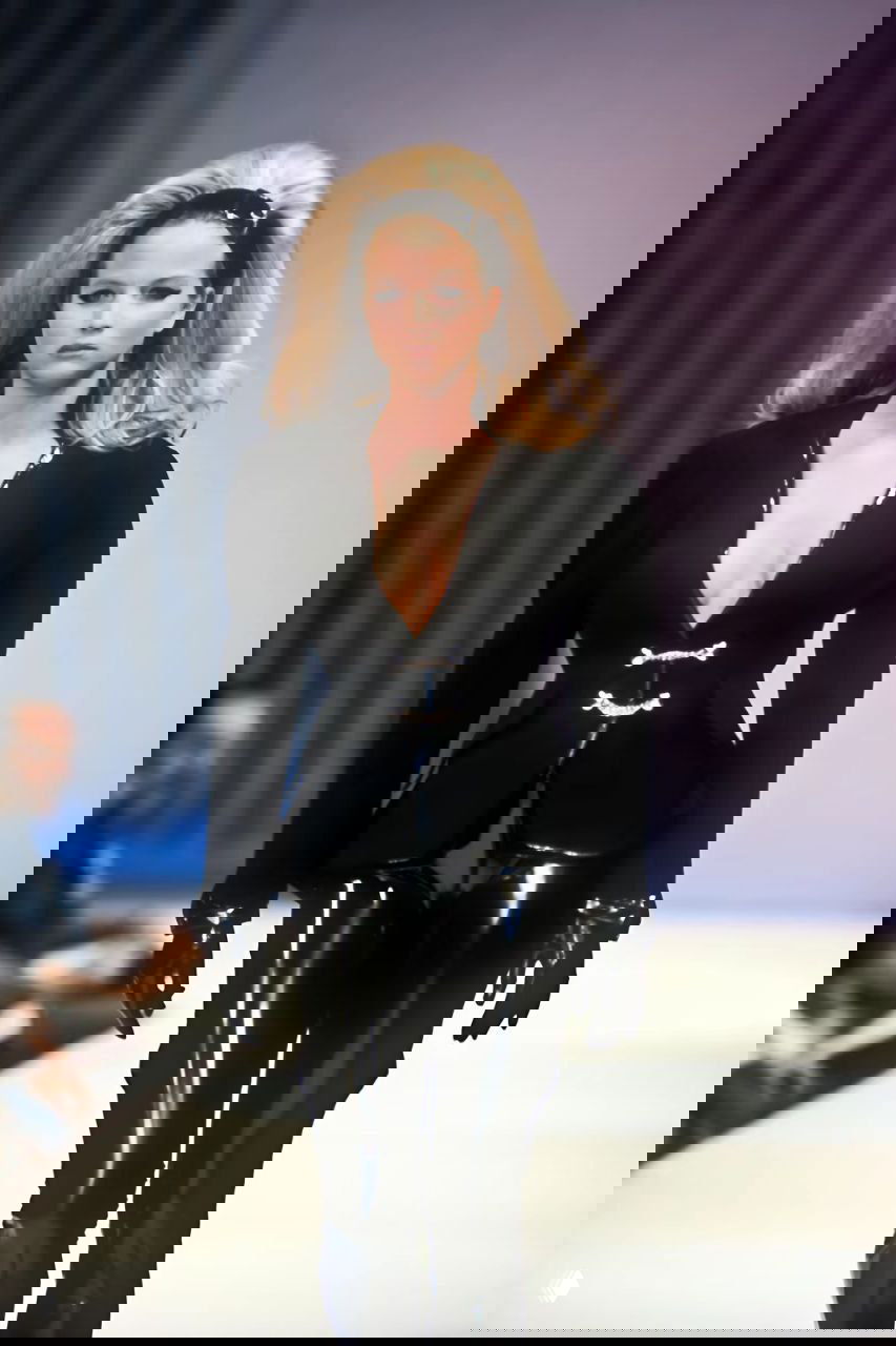 Фигура в чёрном приталенном жакете и лакированных брюках идёт по подиуму — классический силуэт и глянцевые материалы в коллекции Mugler 1990.