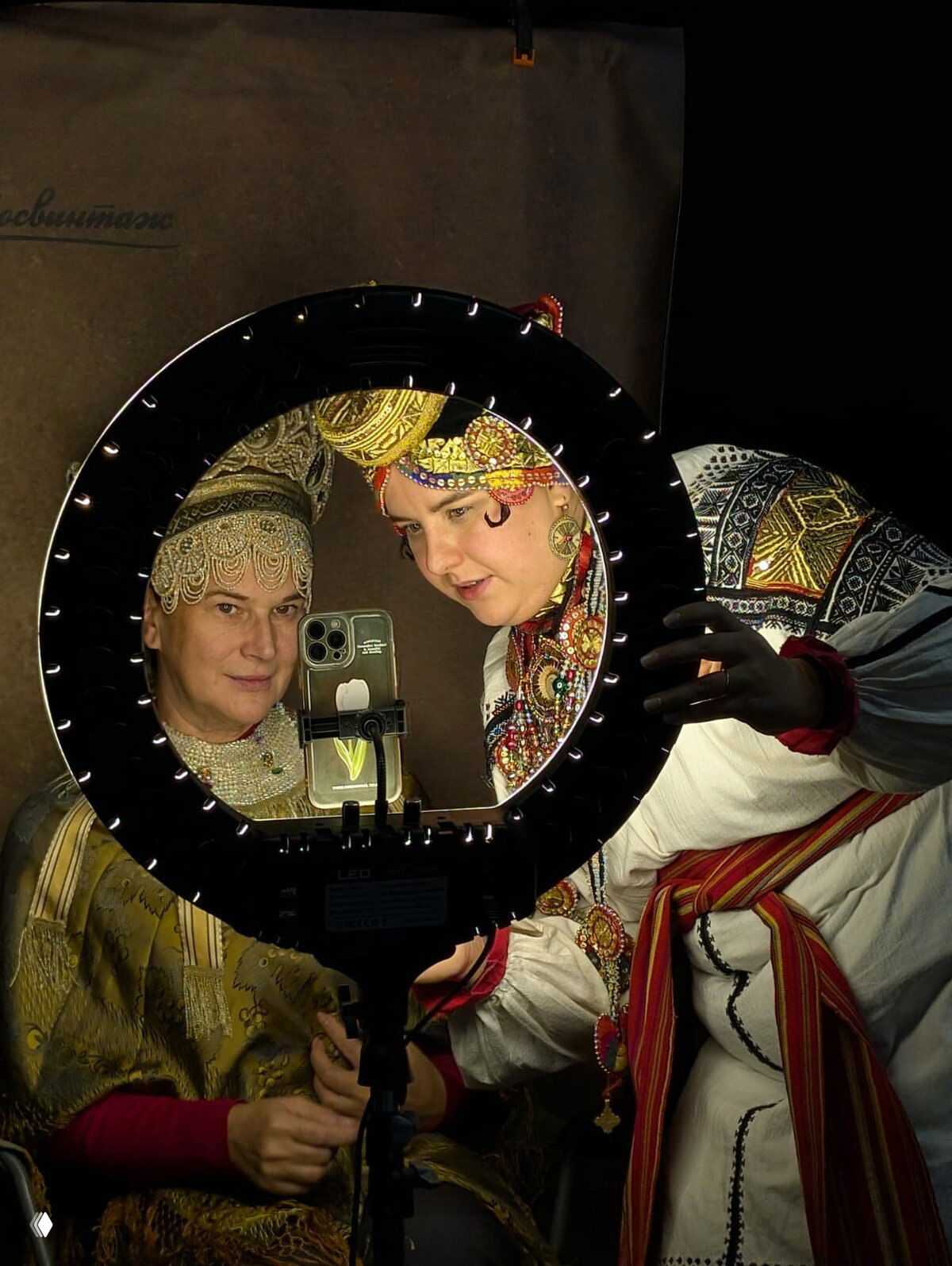 Две женщины в традиционных костюмах смотрят в зеркало через кольцевую лампу на фотозоне выставки; атмосфера мероприятия и костюмы на переднем плане.