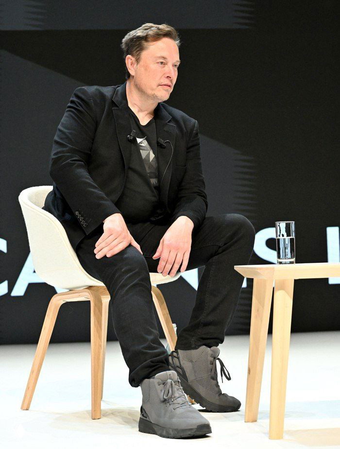 Мужчина в деловом костюме сидит на сцене во время публичного выступления под прожекторами; кадр показывает обувь, стул и стакан воды на столике рядом.