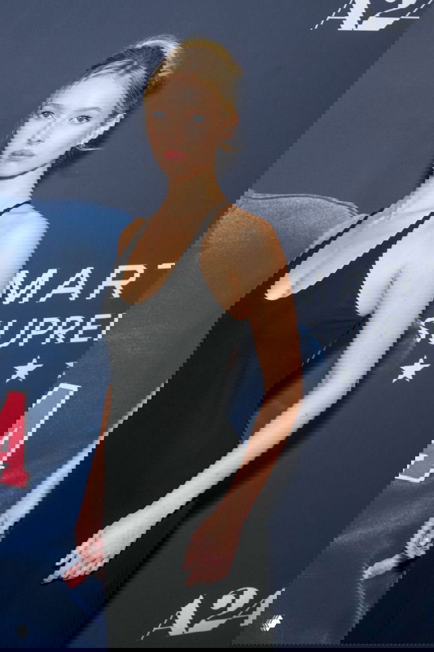 Женщина в минималистичном чёрном платье Calvin Klein на премьере фильма, полный рост на тёмном фоне баннера и красной дорожки