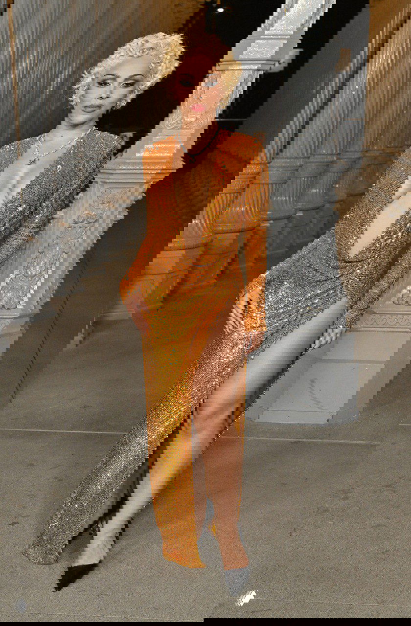 Женщина в золотистом блестящем платье с глубоким декольте и высоким разрезом стоит у колонны музейной инсталляции, вечерний свет.