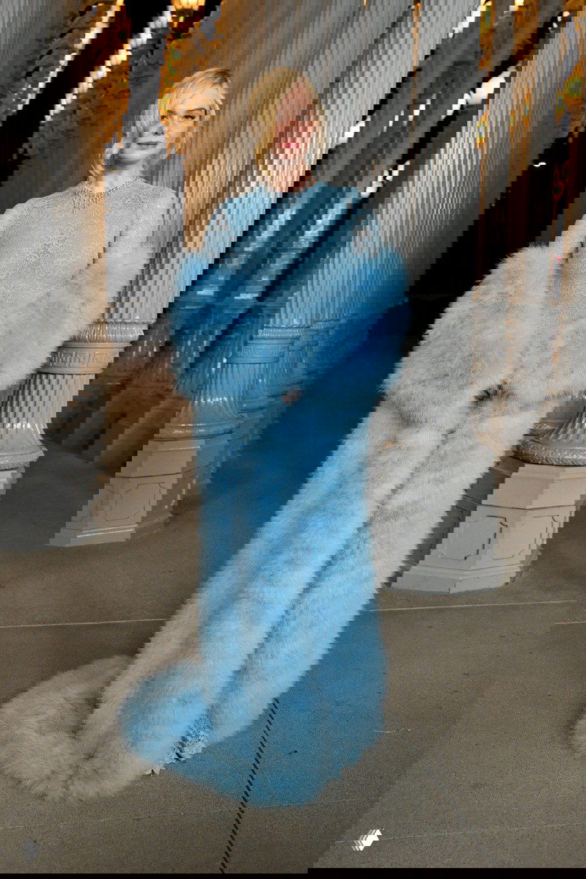 Женщина в голубом пушистом длинном платье с шубоподобным шлейфом позирует на дорожке между колоннами музейной инсталляции вечером.