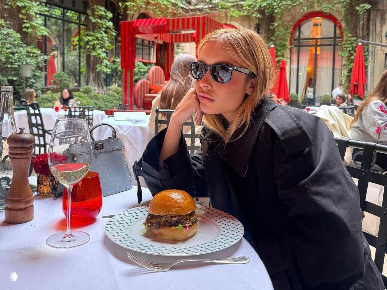 Женщина в солнечных очках и чёрном пальто сидит в стильном уличном кафе за сервированным столом с бургером на тарелке и бокалом вина, городской фон.