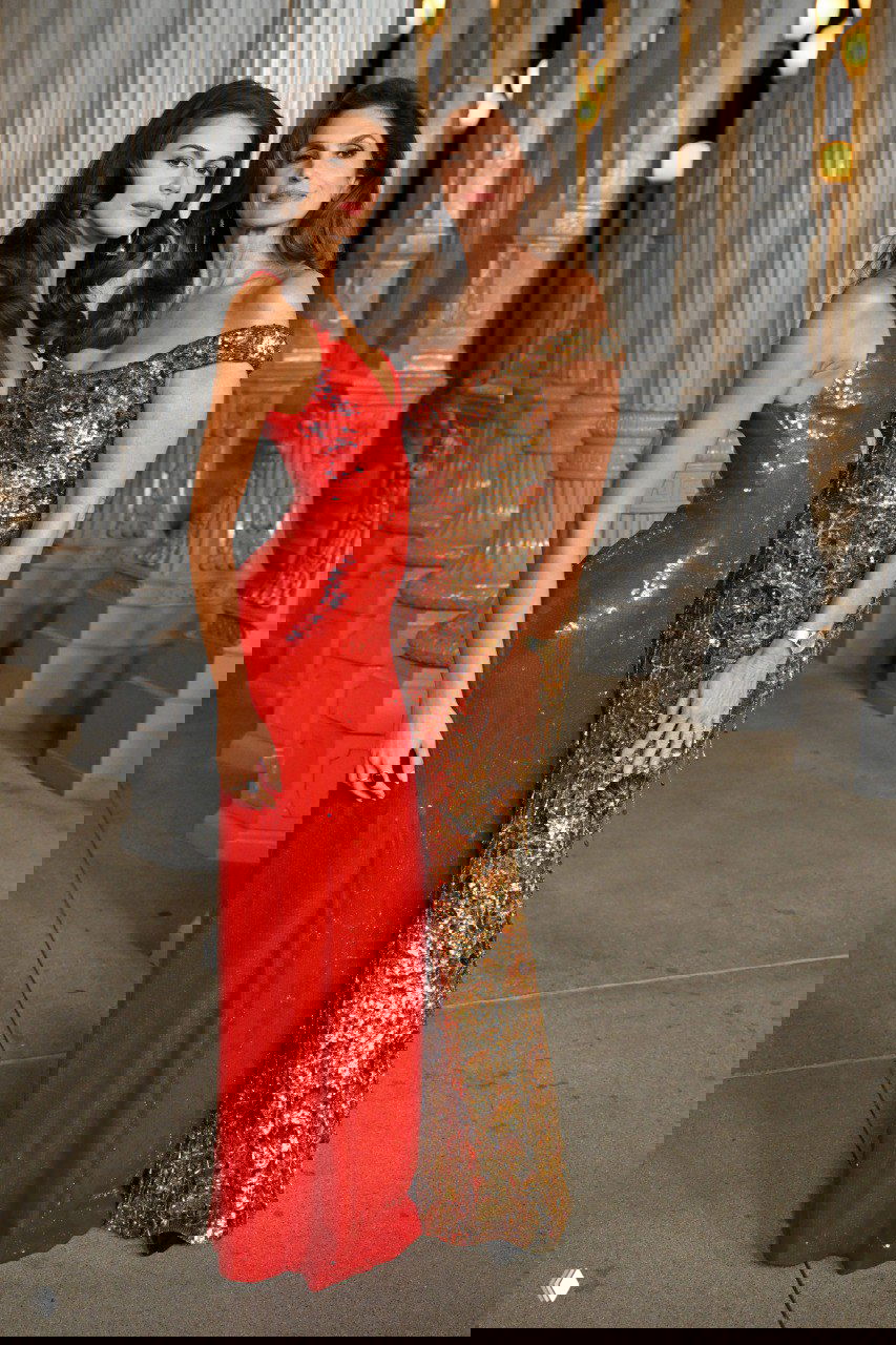 Две женщины в длинных вечерних платьях — красном и золотистом — позируют вместе у колонн музейной инсталляции, полный рост.