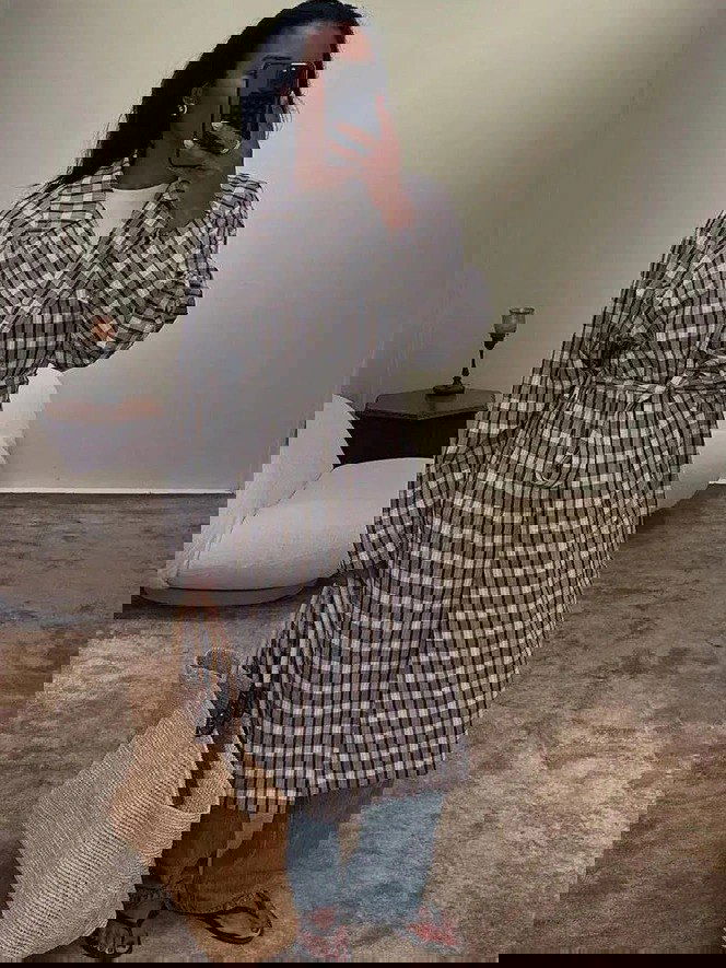 Селфи в зеркале: женщина в длинном клетчатом плаще с поясом, джинсы и соломенная сумка — базовый городской лук с сдержанной клеткой для прохладной погоды.