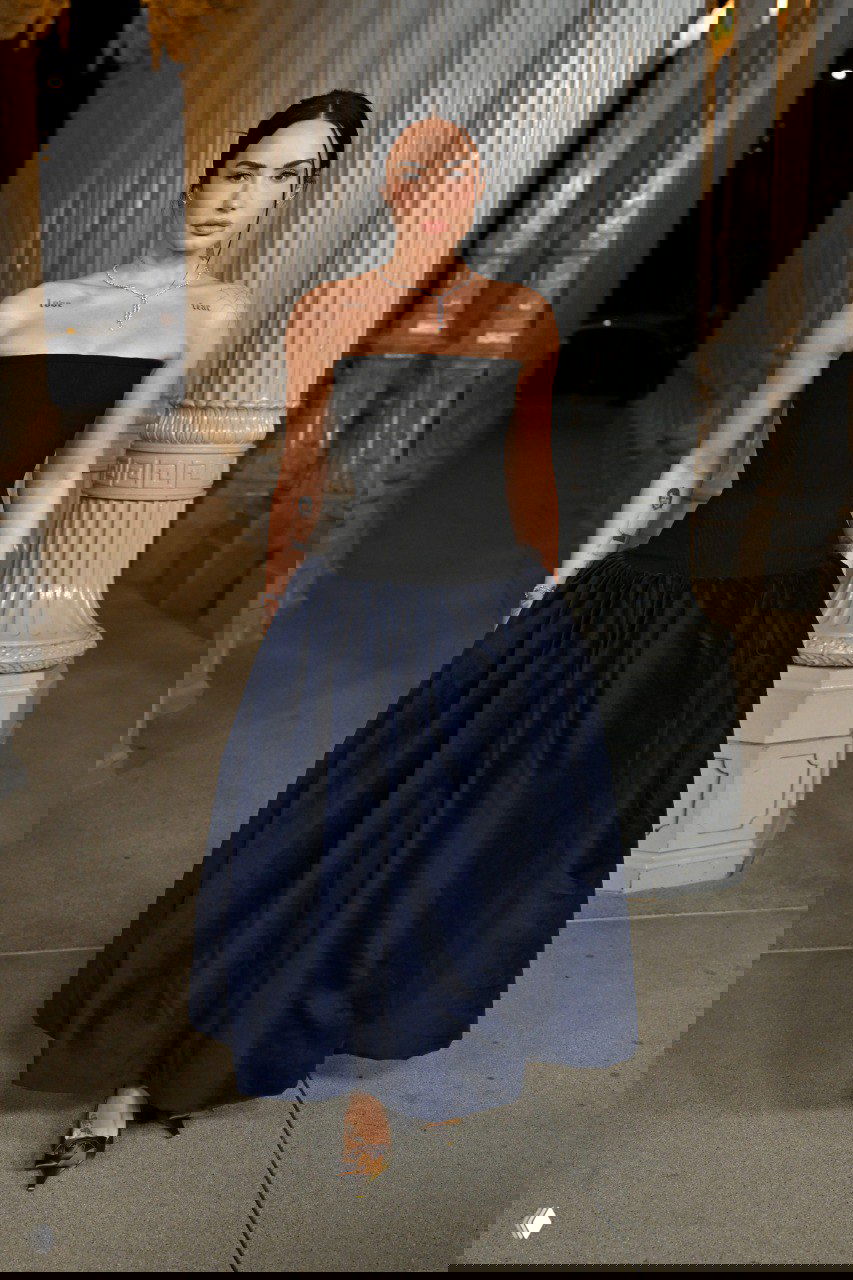 Женщина в темном корсетном лифе и пышной юбке темно-синего тона стоит на дорожке между колоннами — сдержанный вечерний образ.