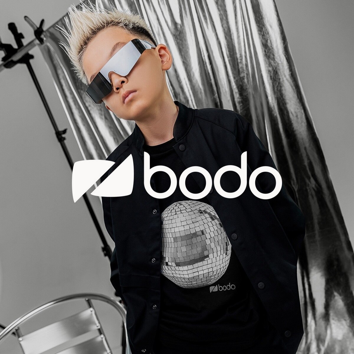 Модель ребёнок в тёмной одежде и очках на студийном фоне с логотипом «bodo», создаёт взрослый образ в детской моде.