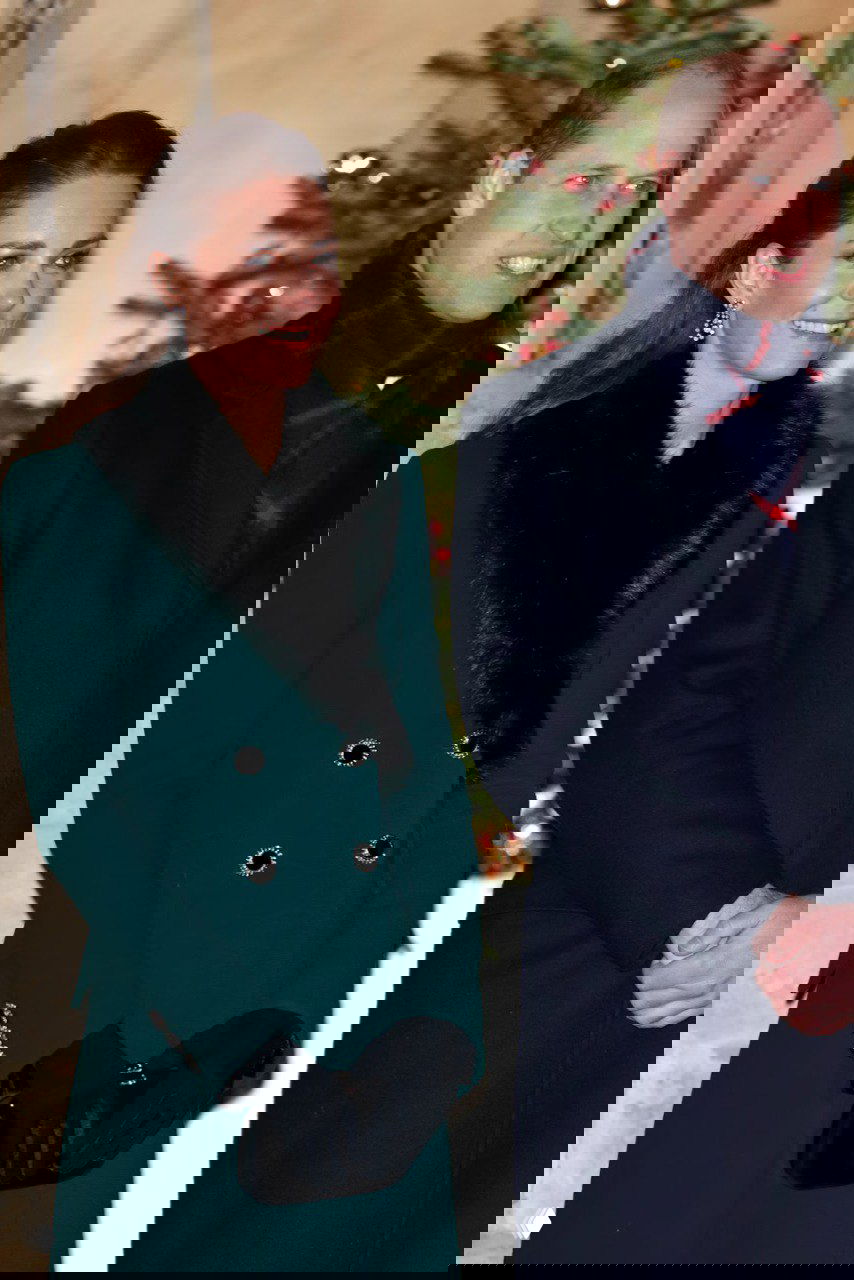 Пара в тёмных пальто стоит на фоне праздничной ёлки: образ с тёмным верхним платьем и классическим пальто у входа в церемонию.