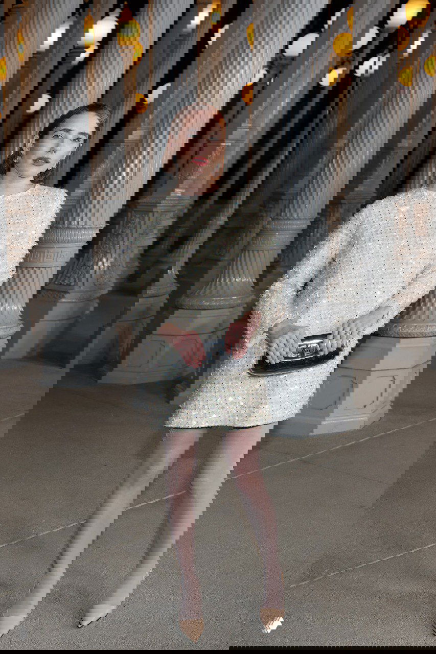 Женщина в коротком светлом платье с текстурой и клатчем стоит у колонны — элегантный минималистичный образ в светлых тонах.