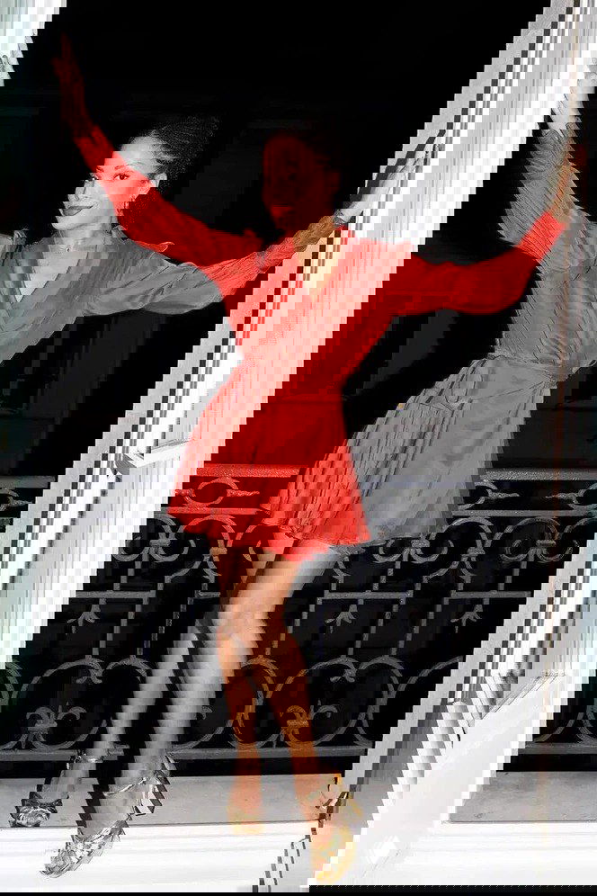 Женщина в красном коротком платье стоит на балконе в золотых босоножках на каблуке — праздничный образ для новогодней вечеринки.