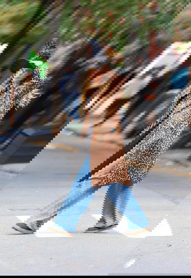 Кадр на улице: женщина в длинном леопардовом пальто и широких джинсах переходит зебру, городской фон и прохожие на заднем плане.