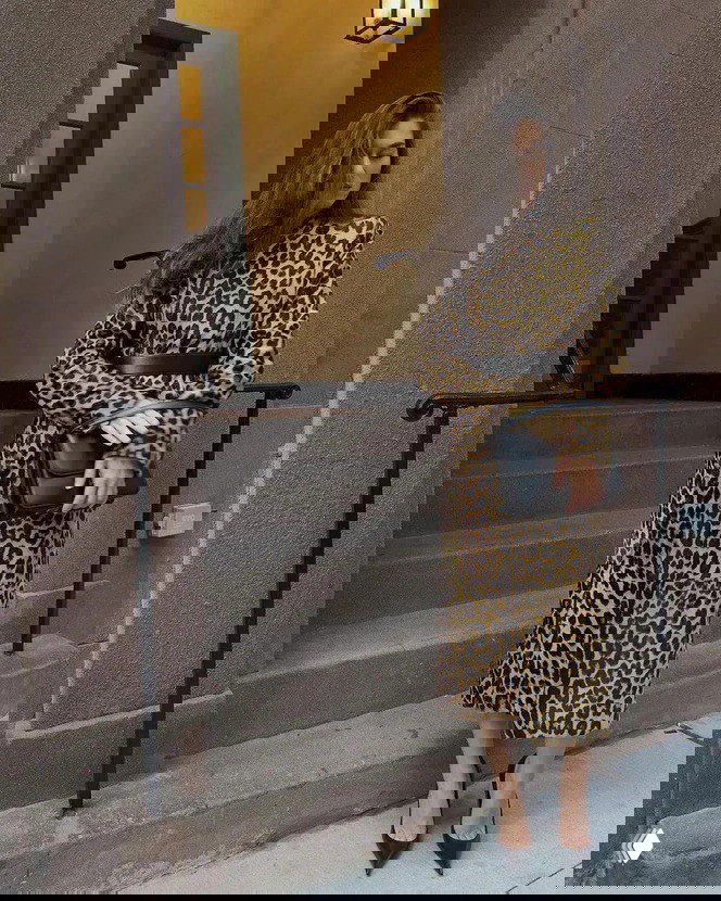 Стильный кадр у ступеней: женщина в длинном пальто с леопардовым принтом, с поясом и кожаной сумкой позирует у входа городского дома.