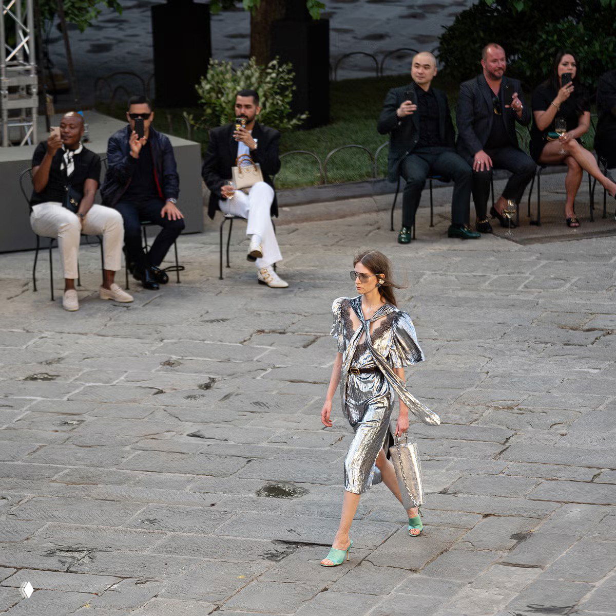 Модель в серебристом платье идёт по открытому каменному подиуму во время показа Gucci; на заднем плане сидящие зрители и уличная локация показа мод