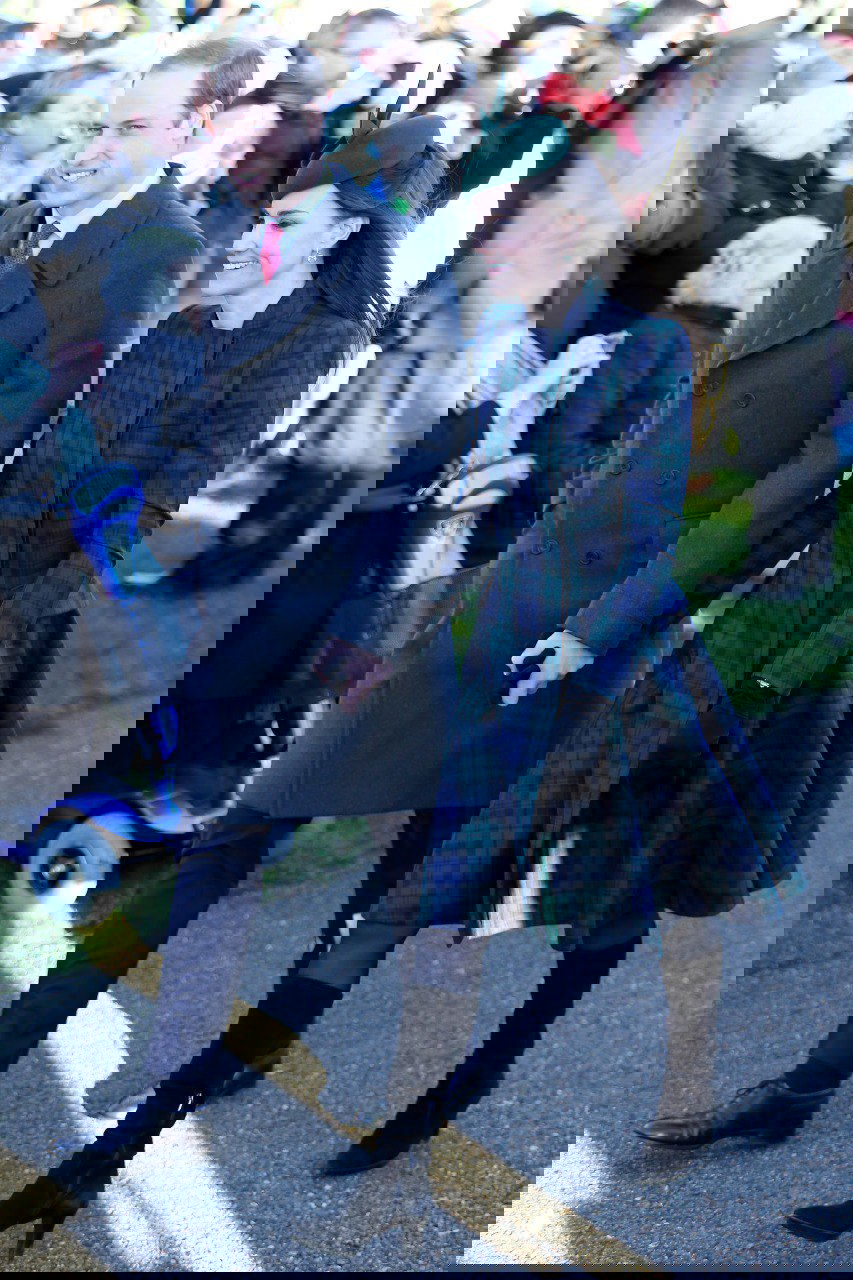 Пара идёт по тропинке среди зрителей: женщина в тёмно-зелёном клетчатом пальто и мужчина в тёмном пальто на фоне толпы и природы.