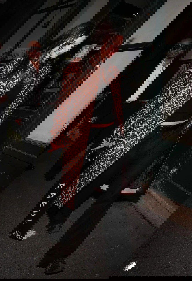 Ночной уличный снимок: женщина в леопардовом пальто и чёрном наряде идёт по тротуару возле входа в заведение, вокруг охранник и прохожие.