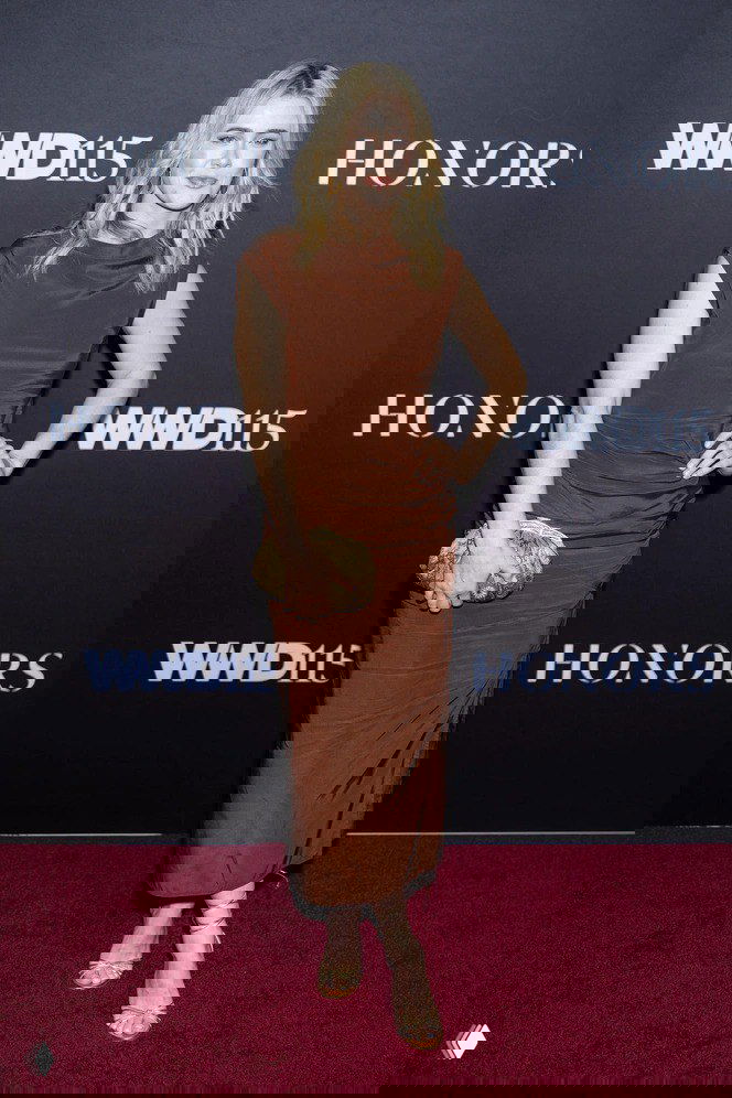 Блондинка на красной дорожке в длинном коричневом платье и золотистых открытых туфлях с ремешками — классический наряд для событий.