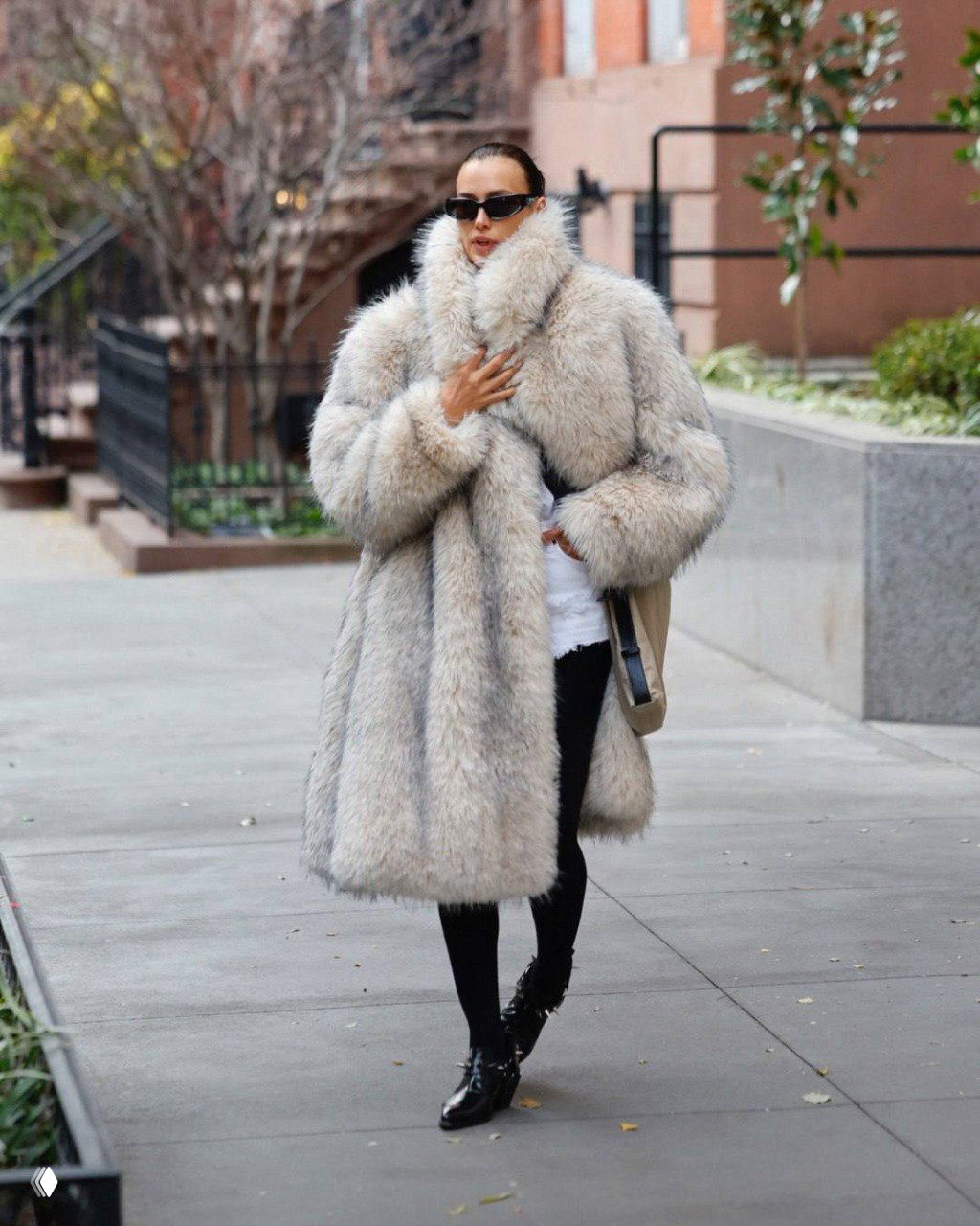 Женщина в крупной объёмной белой шубе и тёмных солнцезащитных очках идёт по улицам Нью‑Йорка в зимнем городском аутфите.