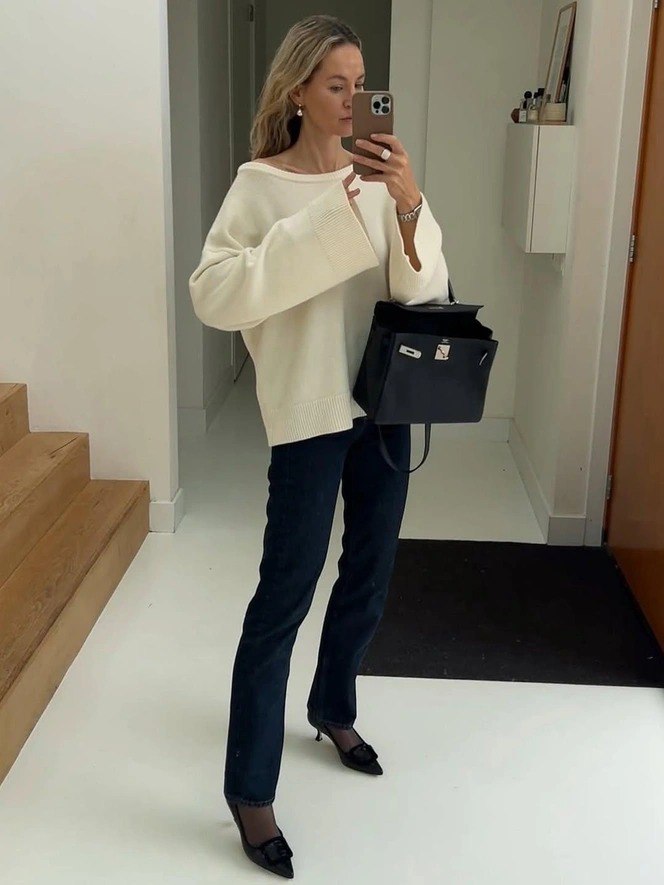 Женщина в светлом объёмном трикотаже и тёмных джинсах демонстрирует сочетание фактур и аккуратную обувь на нейтральном фоне интерьера.