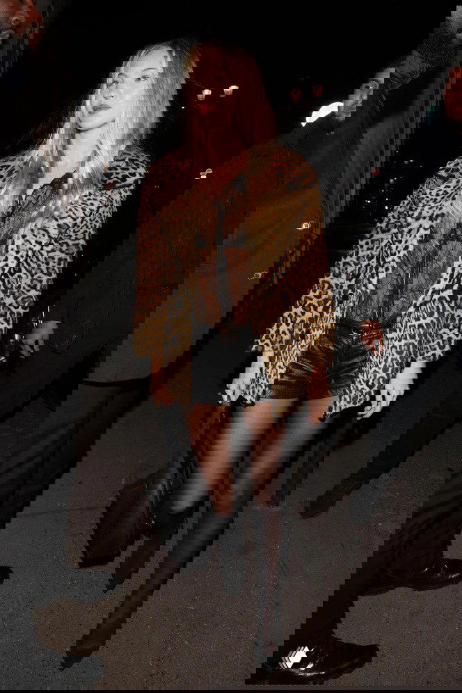 Папарацци-снимок: женщина в коротком леопардовом пальто, прозрачной блузе и шортах выходит вечером из заведения, уличное освещение.