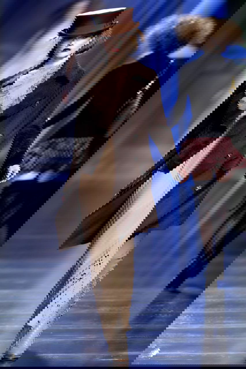 Дефиле Dior осень‑зима 2010: модель в коричневом кожаном плаще и цилиндрической шляпе, образ в викторианской эстетике с современным акцентом.