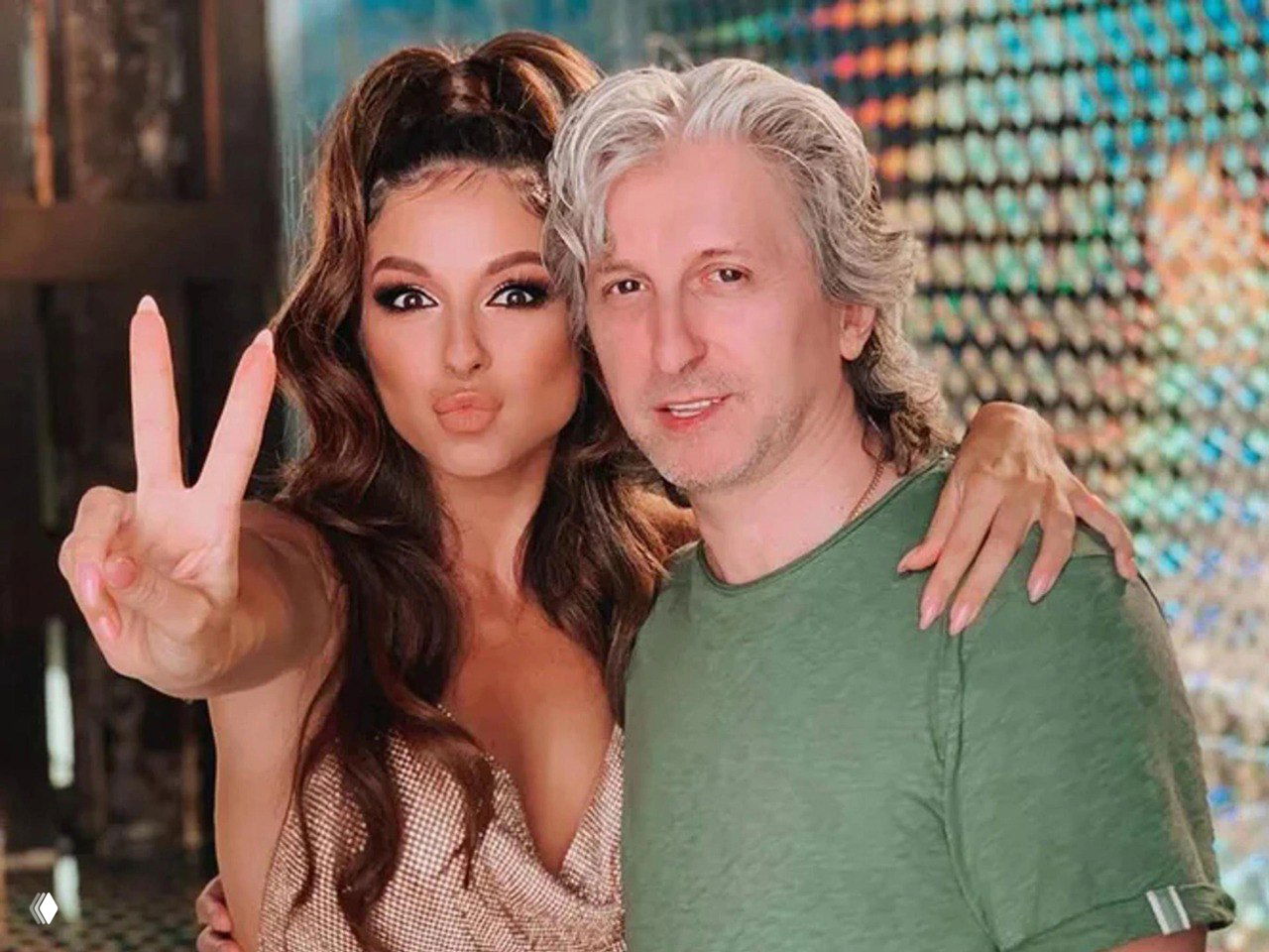 Нюша и Владимир Шурочкин в более неформальной обстановке: певица показывает жест «V», они обнимаются на фоне разноцветной подсветки, улыбаясь в камеру.