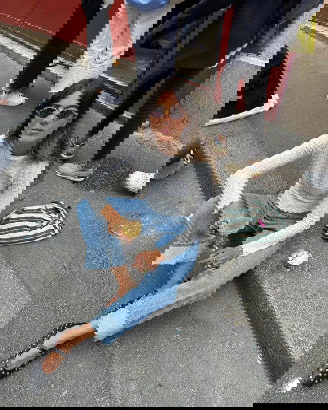 Женщина в джинсах и черных балетках с украшениями сидит на асфальте с бокалом — образ в городском стиле для расслабленного празднования.