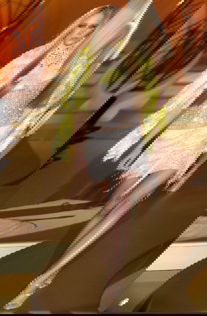 Модель в прозрачном блестящем топе и мини-юбке Versace 2004, на плечах жёлтое перьевое накидное, снимок с подиумного показа на золотом фоне.