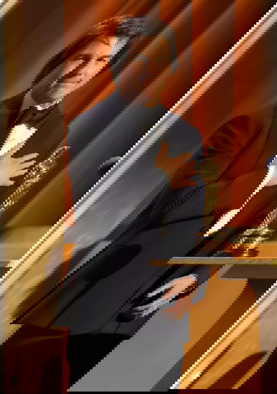 Том Круз в чёрном смокинге на сцене Governors Awards держит статуэтку «Оскар», позирует у подиума на фоне золотистых занавесов