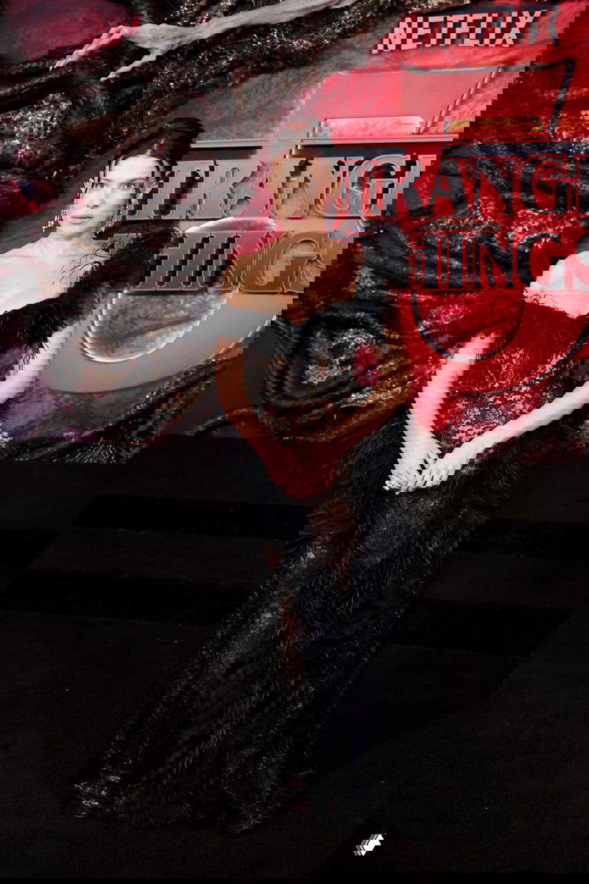 Милли Бобби Браун в полный рост в черном кружевном платье на красной дорожке перед декоративным баннером с логотипом сериала и Netflix